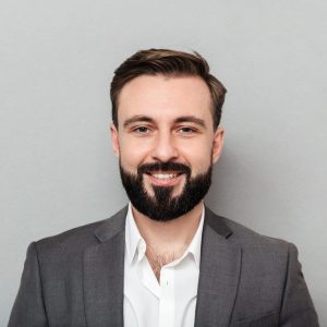 Close up portrait of young bearded man in white shirt and jacket posing on camera with broad smile isolated over gray background